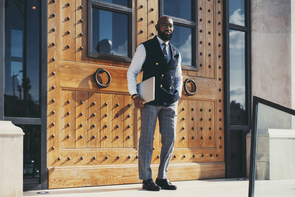 An elegant bald bearded black businessman in glasses and vest standing near huge wooden doors with forged elements after a business meeting, with a laptop in his hand before come downstairs - Фото, изображение