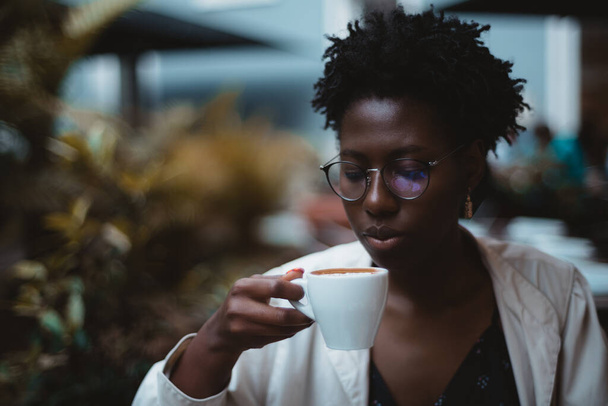 A low-key portrait of a charming young black female with afro curls, in elegant glasses and a white trench coat, drinking a delicious hot chocolate in a dark night cafe; a copy space place on the left - Фото, изображение