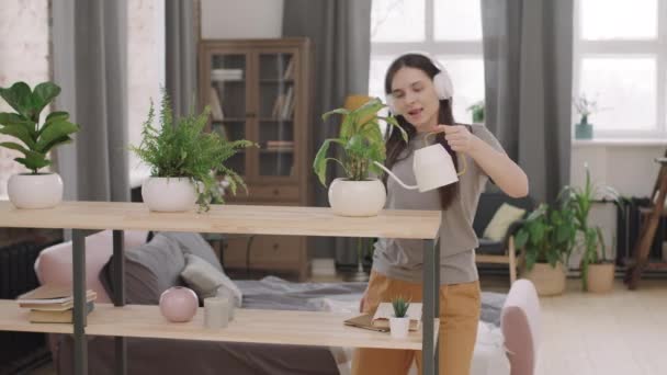 PAN ujęcie młodej kobiety w słuchawkach śpiewającej i tańczącej do muzyki podczas podlewania roślin doniczkowych w jej mieszkaniu - Materiał filmowy, wideo