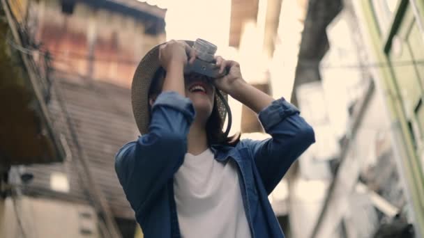 Beautiful woman young Asian traveler wearing retro fedora hat using film camera taking a photo at a small street in Bangkok Thailand. Enjoying traveling on holiday summer. Solo travel concept. - Footage, Video