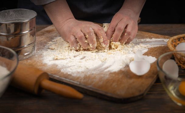 Women's hands, flour and dough. A woman, in an apron cooking dough for homemade baking, a rustic home cozy atmosphere, a dark background with unusual lighting. - Photo, Image