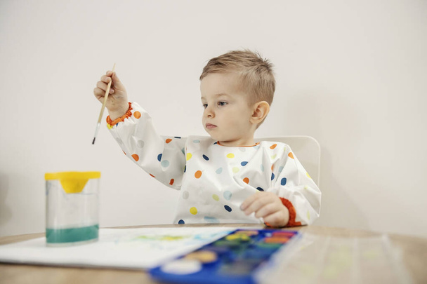 Artist is born. An adorable male toddler dressed in colorful dots sits at a table and draws with a brush and watercolors on paper. Baby boy is focused on drawing. Mental development of the child - Φωτογραφία, εικόνα