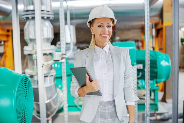 Middle-aged successful blond female supervisor in suit with protective helmet on head standing in power plant with tablet in hands and looking at camera. Elegant woman at work - Photo, Image