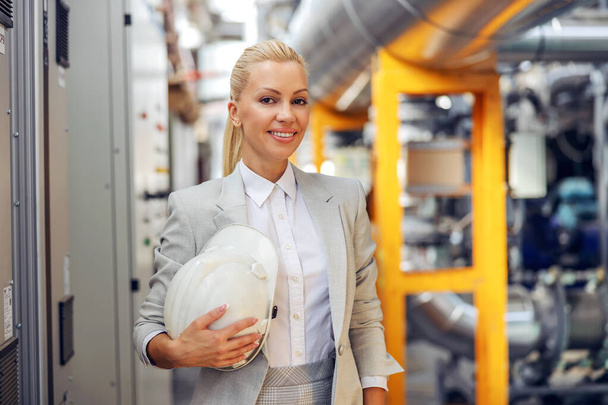 Un retrato de una mujer en un traje elegante en un ambiente de trabajo, mujer CEO. Una supervisora rubia sonriente sosteniendo un casco protector mientras está de pie en una planta de energía. Trabajo en el proyecto - Foto, imagen