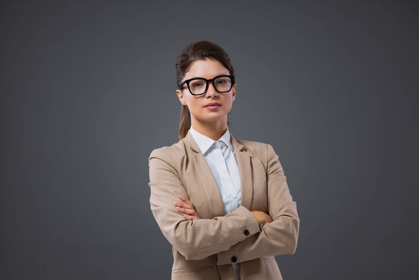 A smart and confident woman at work. Portrait of a serious adult businesswoman standing in front of a gray background with glasses with black eye frames. Professional female boos in elegant wear - Photo, Image
