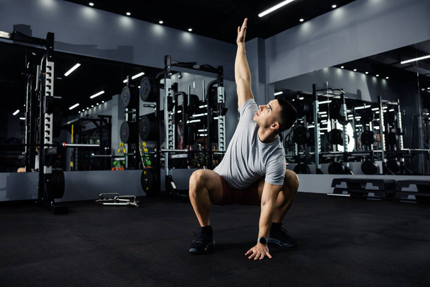 An athletic man in gray sportswear stretches in a dark gym with mirrors. He is in a deep squat position and one arm is extended high above his head. Sport love, healthy lifestyle - Zdjęcie, obraz
