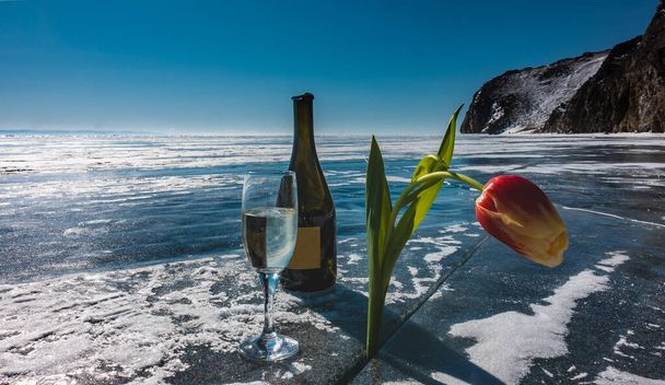 Hay una copa de champán y una botella abierta en el lago congelado. En la grieta de hielo, un brillante tulipán amarillo-rojo inclinó su cabeza. Cordillera contra un cielo azul claro. Baikal. - Foto, imagen