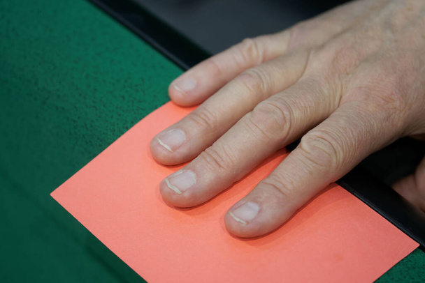 Ein erwachsener männlicher Schiedsrichter oder Wahlteilnehmer zieht einem Karteninhaber eine rote Karte aus dem Papier. Warnung, Verbot oder Ablehnung der Entscheidung. Nahaufnahme. Reportagen. Kein Gesicht - Foto, Bild