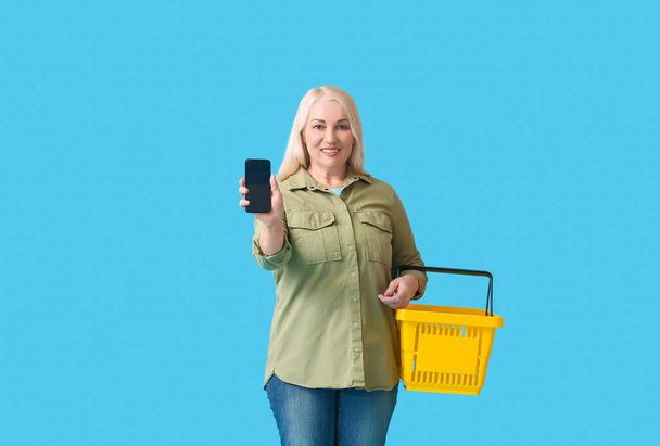Mature woman with shopping basket and mobile phone on color background - Фото, зображення
