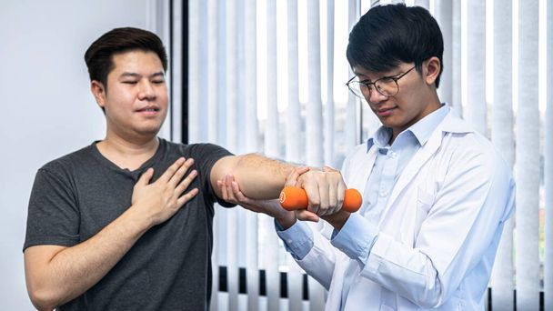 Młody azjatycki fizjoterapeuta pomaga męskiemu pacjentowi rozciągnąć ramię podczas ćwiczeń prawidłowych z hantlami w dłoni podczas treningu ręka do rehabilitacji terapie w klinice - Zdjęcie, obraz