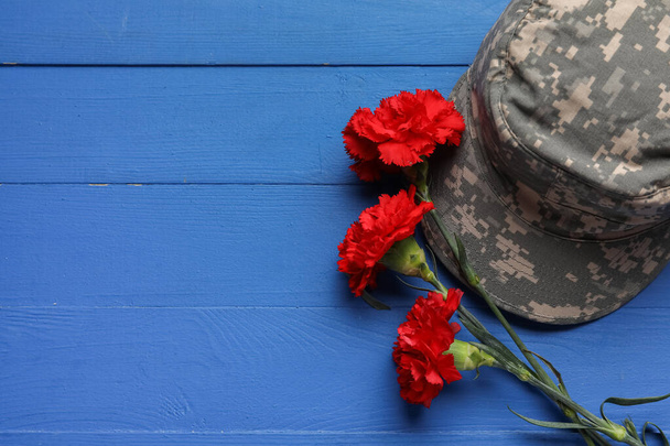 Verse anjer bloemen met militaire pet op kleur houten achtergrond - Foto, afbeelding