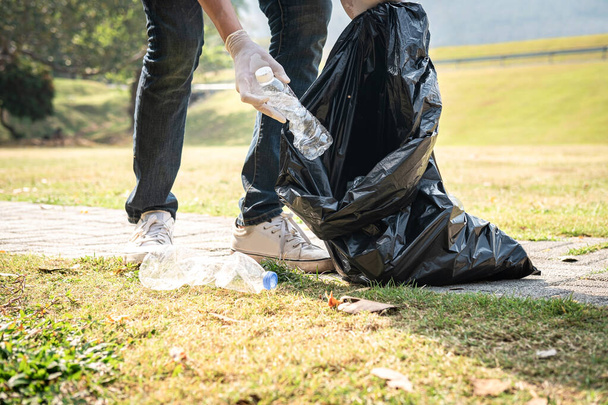 Dobrovolník muž v rukavicích chůze a zastavení sbírat plastové láhve do plastové černé tašky pro čištění parku během činnosti životního prostředí na sběr odpadků - Fotografie, Obrázek
