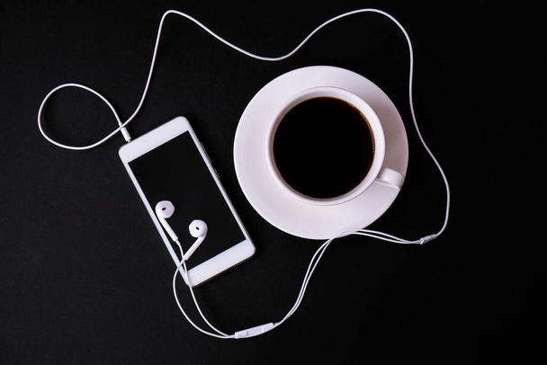 Czarna kawa z telefonem komórkowym i słuchawkami na czarnym tle. Kreatywna koncepcja. Audiobook. Podcast. Odbiór. Monochromatyczne. - Zdjęcie, obraz