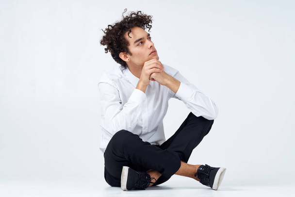 guy with curly hair in a white shirt, sneakers and trousers sits on the floor. High quality photo - Photo, Image
