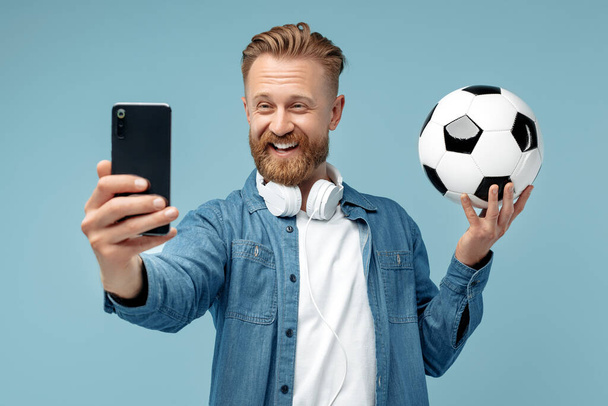 Smiling bearded hipster blonde young man take selfie on smartphone holding classic soccer ball over blue background. - Foto, Imagen