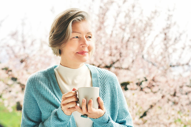 Весенний портрет красивой и элегантной женщины средних лет, позирующей против розового цветущего дерева, держащей чашку кофе или чая - Фото, изображение