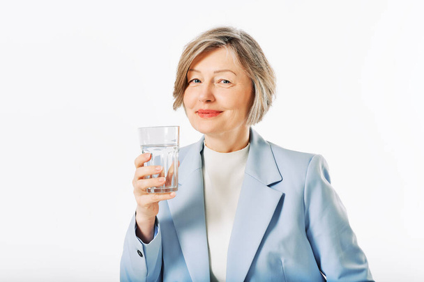 Studio portrét ženy středního věku pózující na bílém pozadí, 55 - 60 let stará modelka držící sklenici vody - Fotografie, Obrázek