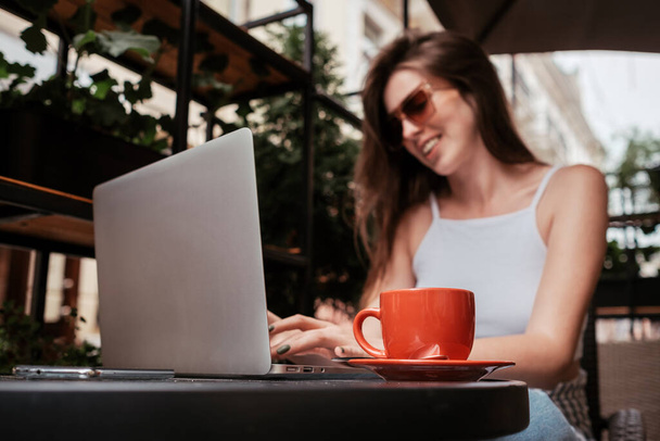 Jovem mulher alegre bebendo e trabalhando em laptop em um café ao ar livre, foco na xícara de café, conceito freelance - Foto, Imagem