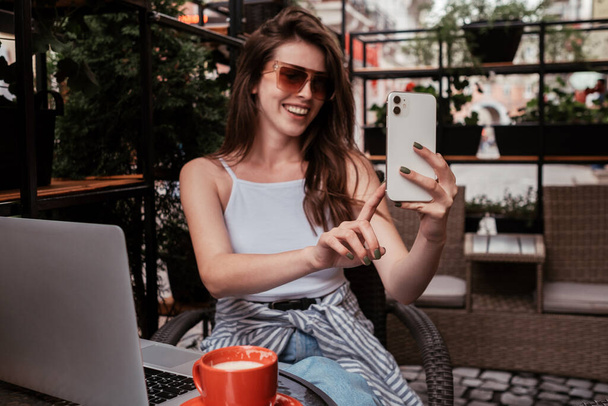 Nuori iloinen nainen käyttää älypuhelinta videopuheluun istuessaan kahvilassa ulkona - Valokuva, kuva