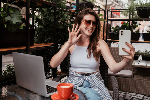 Jonge vrolijke vrouw met behulp van smartphone voor video bellen tijdens het zitten in een café Buiten - Foto, afbeelding