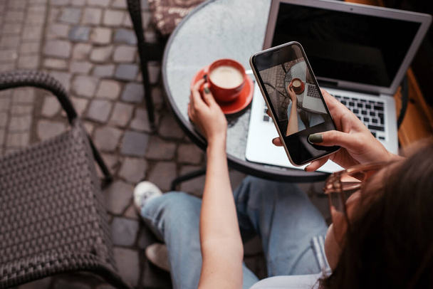 若いブルネットの女性がカフェの屋外に座っている間にスマートフォンでコーヒーの写真を撮る - 写真・画像