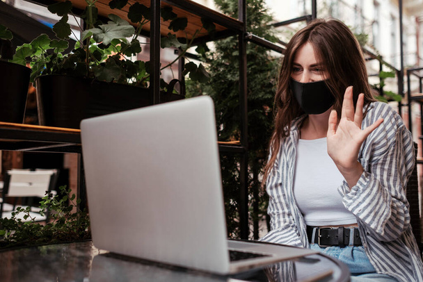 Jeune femme brune portant un masque protecteur pour le visage à l'aide d'un ordinateur portable pour un appel vidéo assis dans un café à l'extérieur, concept de pandémie Covid-19 - Photo, image
