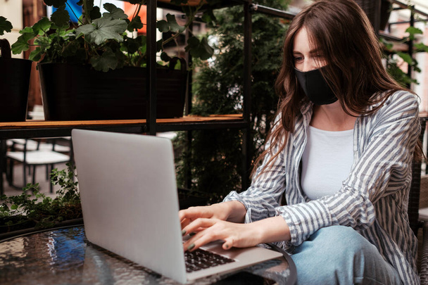 Nuori brunette nainen yllään suojaava kasvot naamio Työskentely kannettavan tietokoneen istuessaan kahvilassa ulkona, pandemia Covid-19 Concept - Valokuva, kuva