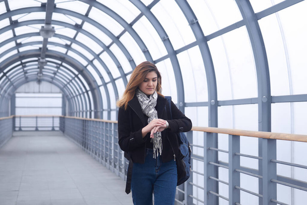 young woman walks along a covered pedestrian overpass and looks at her watch - Foto, imagen