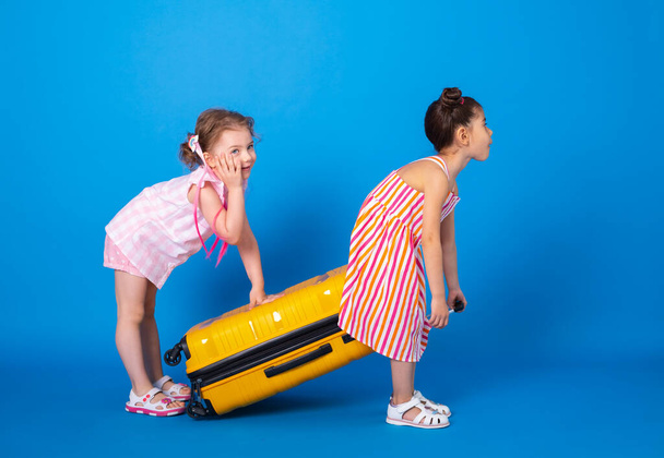 funny little girls friends in colorful clothes carrying yellow suitcase on blue background. - Photo, Image
