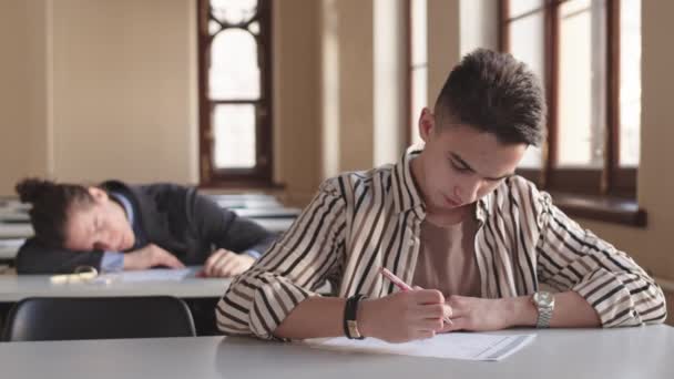 Sınav yapan karışık ırktan erkek öğrenci, sınıfta kalemle yazı yazan, arkasında okul sıralarında uyuyan bulanık sınıf arkadaşı. - Video, Çekim