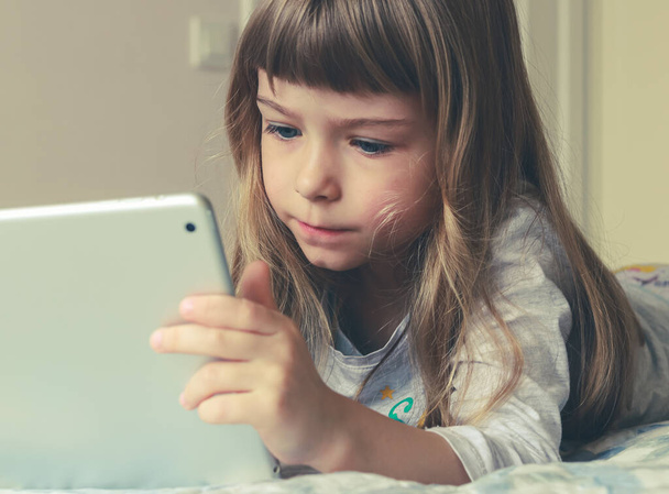 Uzun sarı saçlı bir kız yatağa uzanıyor ve tablet ekranına bakıyor. - Fotoğraf, Görsel