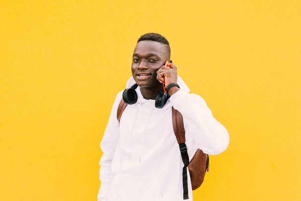 Młody, afrykański czarny mężczyzna rozmawiający ze smartfonem ze słuchawkami bezprzewodowymi patrzącymi w kamerę. Żółte tło ściany, w białej bluzie i plecaku - Zdjęcie, obraz