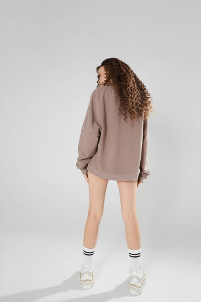 A young model woman with perfect long legs and slim body wearing a grey sweatshirt and trendy sneakers posing with her back at the white background, isolated with copy space - Photo, Image