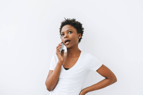 Emotional beautiful young African American woman in white T-shirt using mobile phone isolated on white background - Fotoğraf, Görsel