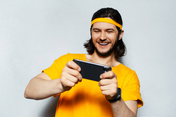Portrait of young excited man using smartphone, wearing yellow shirt on background of grey textured wall. - Foto, imagen