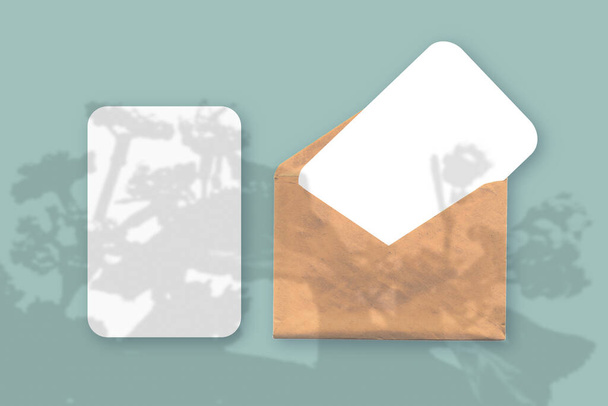 Makieta z nakładką cieni roślinnych na kopertę z dwoma arkuszami teksturowanego białego papieru na zielonym tle stołu. Orientacja pozioma - Zdjęcie, obraz