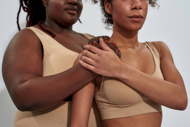 Foto recortada de dos mujeres afroamericanas en ropa interior beige apoyándose unas a otras, de pie juntas aisladas sobre fondo gris - Foto, Imagen