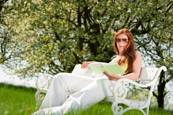 Mujer pelirroja leyendo libro en banco blanco en prado verde, DOF poco profundo
 - Foto, Imagen