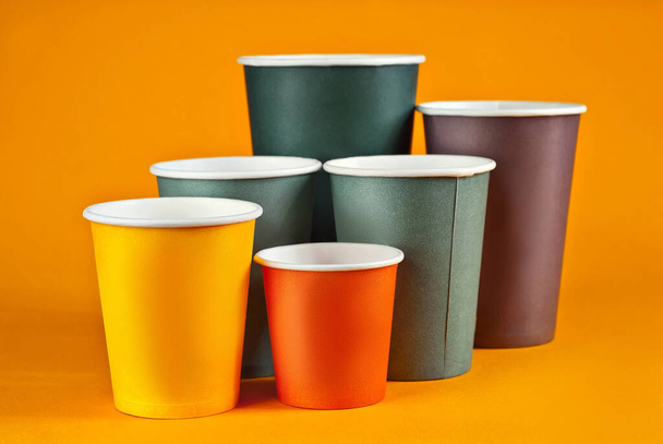 Disposable paper cups on orange background. Glasses of different sizes and colors close up. Party utensils for different drinks. - 写真・画像