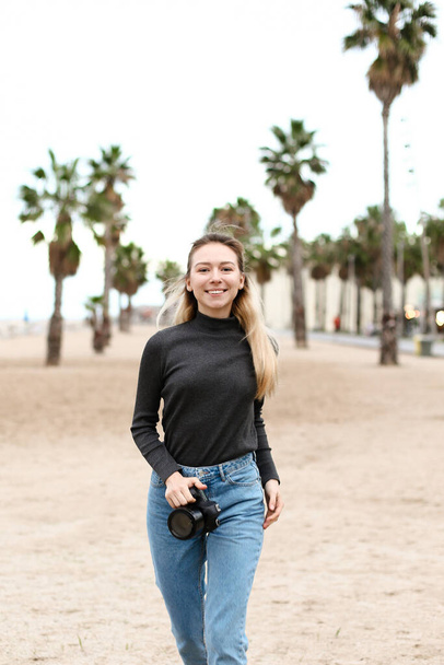 Молодая женщина идет по пляжу с камерой и в черной блузке. - Фото, изображение