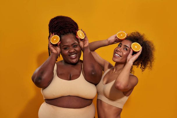 Δύο παιχνιδιάρες αφροαμερικανίδες που διασκεδάζουν, κρατώντας το μισό ώριμο ζουμερό πορτοκάλι, ποζάροντας απομονωμένες σε πορτοκαλί φόντο. - Φωτογραφία, εικόνα