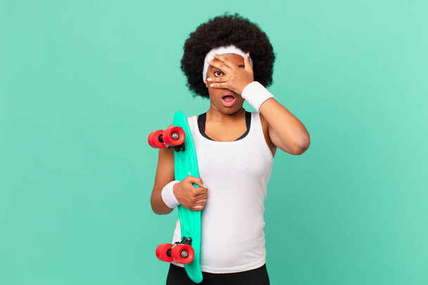 afro woman looking shocked, scared or terrified, covering face with hand and peeking between fingers. skateboard concept - 写真・画像