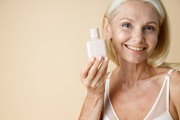 Close up portrait of gorgeous mature blonde woman in underwear smiling at camera, holding white bottle of cream or lotion for skin care, posing isolated over beige background - Foto, imagen