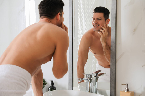 Handsome man touching his smooth face after shaving near mirror in bathroom - Фото, изображение