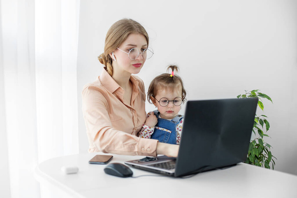 Młoda kobieta pracuje w domu używając laptopa. Małe dziecko siedzi w ramionach matki. Praca na urlopie macierzyńskim. - Zdjęcie, obraz