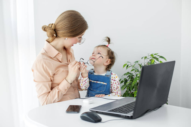 Onze moeder werkt thuis terwijl ze tijd doorbrengt met haar dochter. Jonge vrouw werkt thuis met een laptop. - Foto, afbeelding