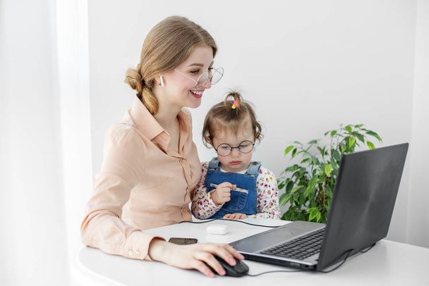 Zajęta matka i dziecko w domu pracują nad komputerem. Kobieta się uśmiecha. Praca na urlopie macierzyńskim. Mała dziewczynka bawi się blisko mamy. - Zdjęcie, obraz