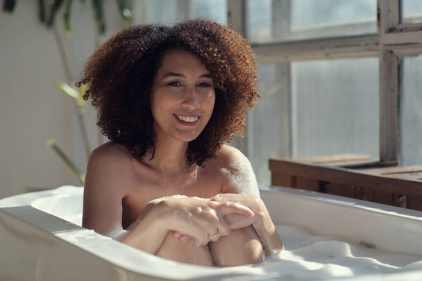 Mujer afroamericana sonriente y relajante bañándose en una bañera llena de espuma. Un tiempo increíble. estilo de vida concepto de personas - Foto, imagen