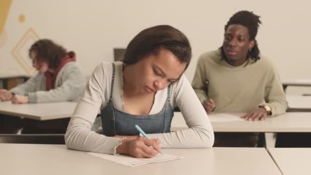 Pecho-up de estudiante de raza mixta, sentada en el escritorio en el aula, escribiendo en el examen, su borrosa compañera de clase africana espiando en su sábana detrás de la espalda - Imágenes, Vídeo