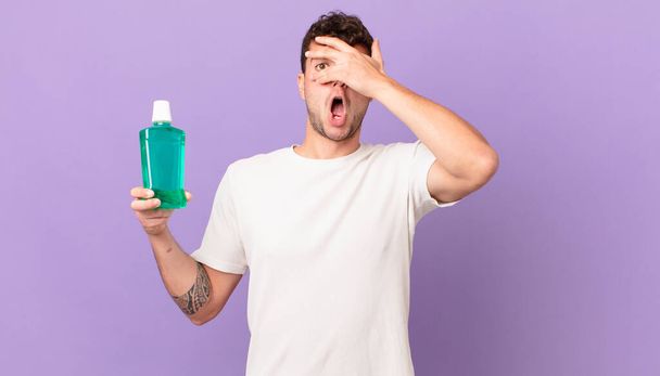 man with mouthwash looking shocked, scared or terrified, covering face with hand and peeking between fingers - Foto, Imagem